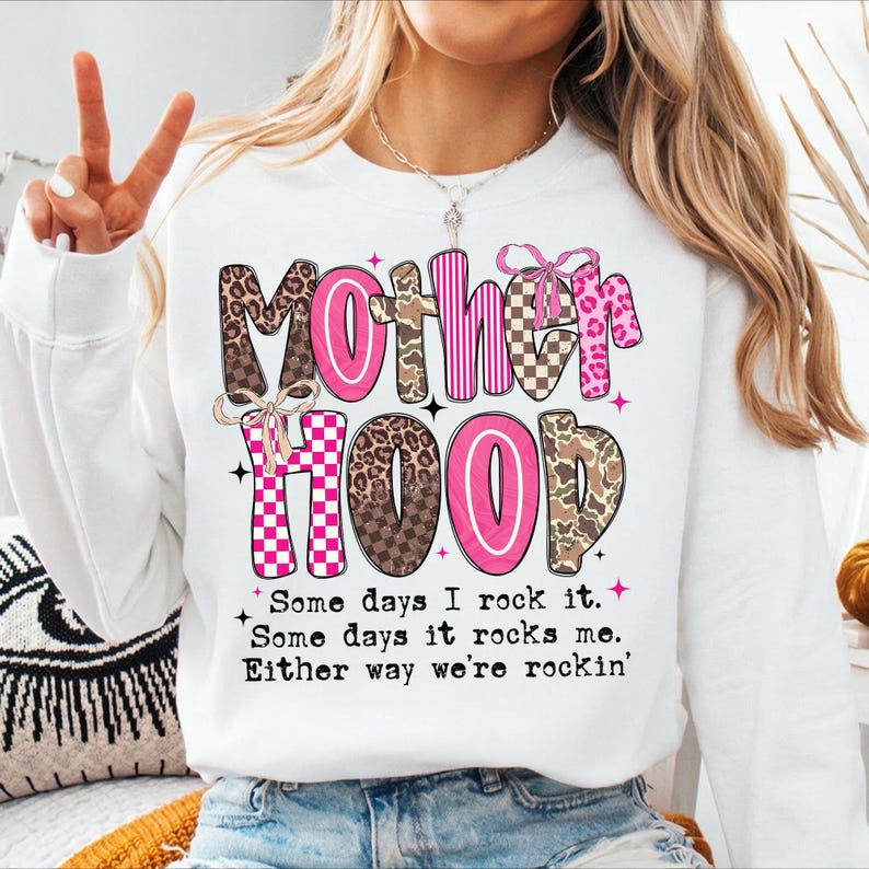 Puede incluir: Sudadera blanca con la palabra "Motherhood" en rosa y letras estampadas. El texto "Some days I rock it. Some days it rocks me. Either way we're rockin'" est&aacute; impreso debajo.