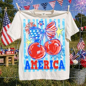 May include: Cream-colored t-shirt with a patriotic design. The graphic features two red cherries with a blue and white bow, fireworks, and the word "AMERICA" in blue and red. The shirt is hanging on a clothesline with American flags in the background.