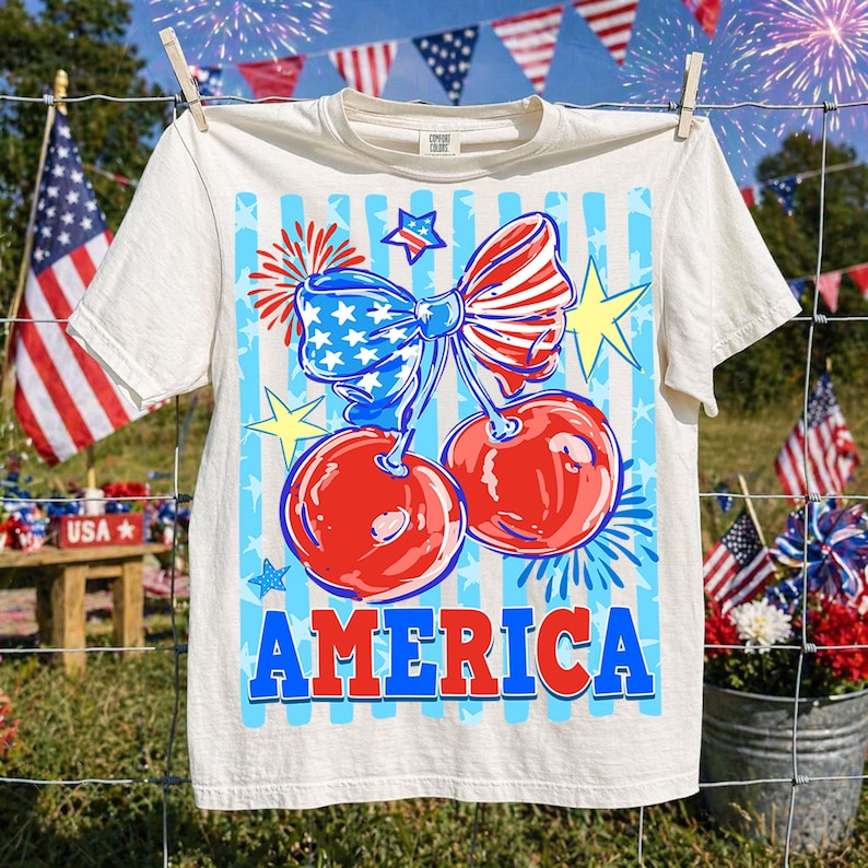 May include: Cream-colored t-shirt with a patriotic design. The graphic features two red cherries with a blue and white bow, fireworks, and the word "AMERICA" in blue and red. The shirt is hanging on a clothesline with American flags in the background.