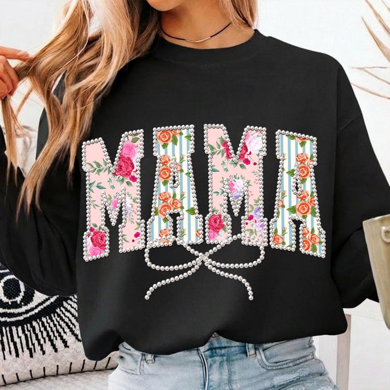 May include: Black sweatshirt with the word "MAMA" in large, floral-filled letters. The letters are outlined with white pearls and feature a pink and blue striped background. A pearl bow is below the letters.