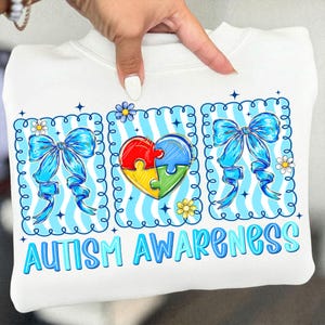 May include: White sweatshirt with a colorful graphic promoting autism awareness. The design includes a puzzle heart, blue bows, and the words "AUTISM AWARENESS" in blue. The sweatshirt is held up by a hand.