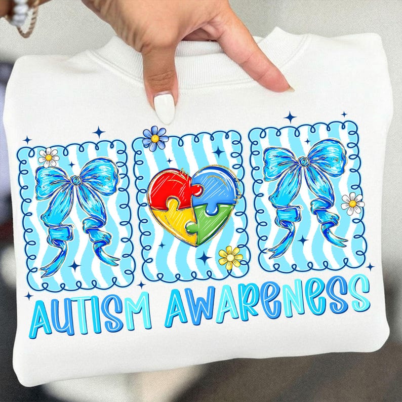 May include: White sweatshirt with a colorful graphic promoting autism awareness. The design includes a puzzle heart, blue bows, and the words "AUTISM AWARENESS" in blue. The sweatshirt is held up by a hand.