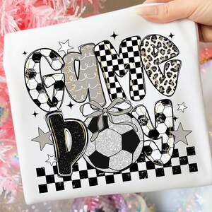 May include: White t-shirt with the text "Game Day" in a playful design. The letters feature soccer ball patterns, leopard print, and checkered patterns. A soccer ball with a bow is in the center, with star accents.