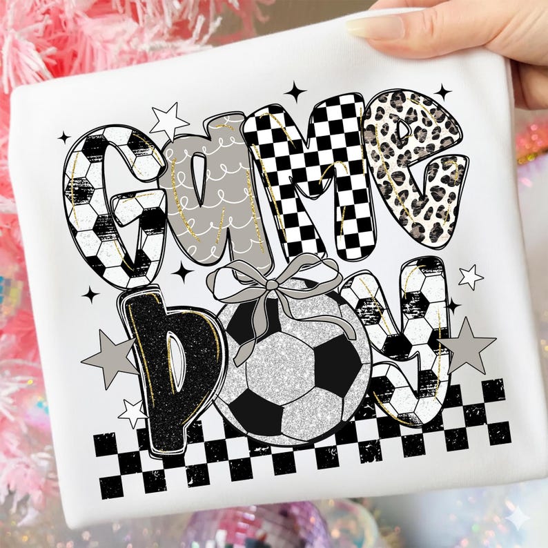 May include: White t-shirt with the text "Game Day" in a playful design. The letters feature soccer ball patterns, leopard print, and checkered patterns. A soccer ball with a bow is in the center, with star accents.