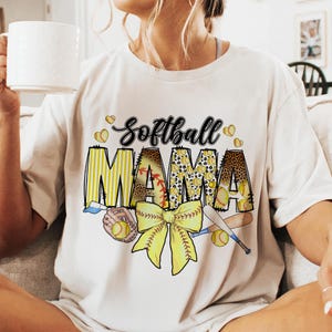 May include: White t-shirt with a graphic design of a softball, a bow, and the text "Softball Mama".