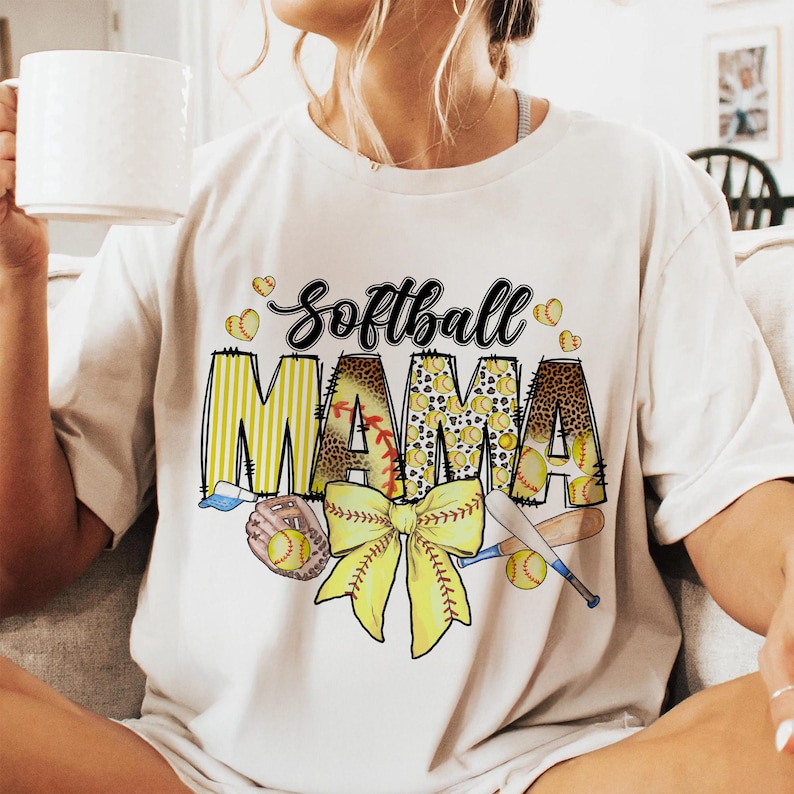 May include: White t-shirt with a graphic design of a softball, a bow, and the text "Softball Mama".
