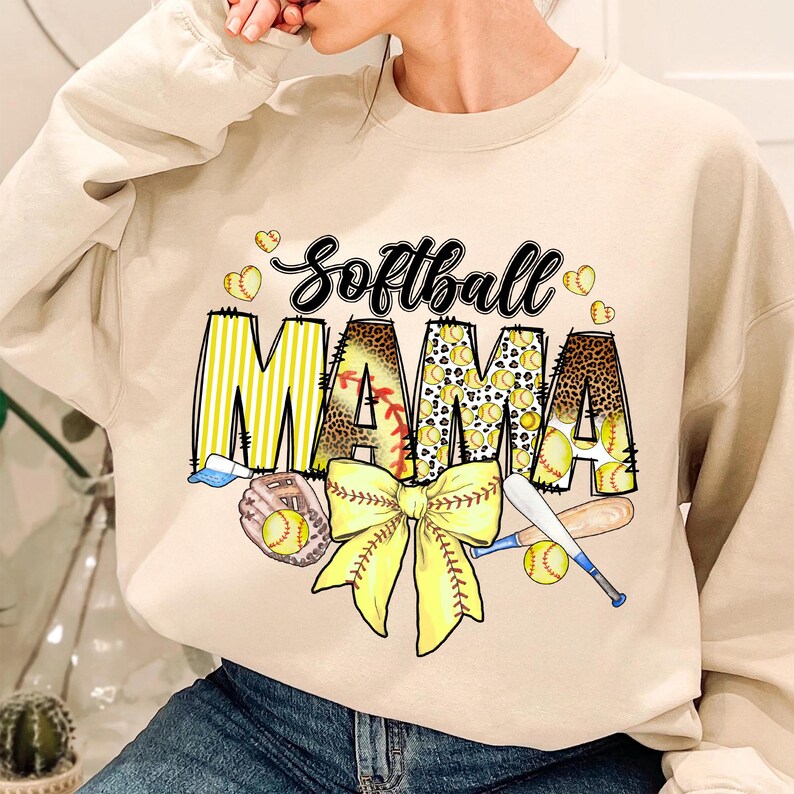 May include: A beige sweatshirt with the text "Softball Mama" in yellow and brown letters. The letters are decorated with softball-themed patterns, including baseballs, bats, and a yellow bow.