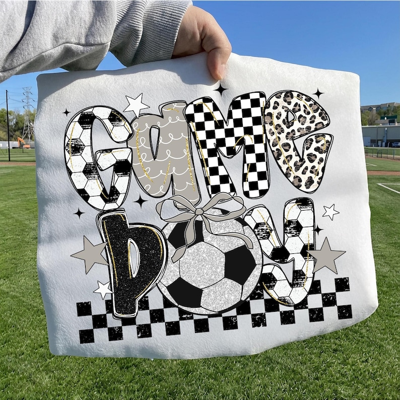 May include: White soccer-themed blanket with the words "Game Day" in a mix of fonts and patterns. The letters feature soccer ball designs, checkered patterns, and leopard print. A soccer ball with a bow is in the center.