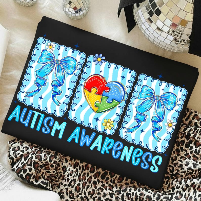 May include: Black t-shirt with the words "AUTISM AWARENESS" in blue. The design includes three blue and white striped squares with a blue bow and a puzzle piece heart. Daisies and stars are also included in the design.