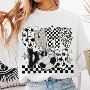 May include: White sweatshirt with the text "Game Day" in a mix of fonts and patterns, including soccer ball designs, a checkered pattern, and leopard print. A soccer ball with a bow is in the center.