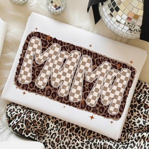 May include: White t-shirt with the word "MAMA" in a brown and tan checkered design, outlined with small silver beads, set against a leopard print background. The shirt is folded, with a leopard print fabric visible.