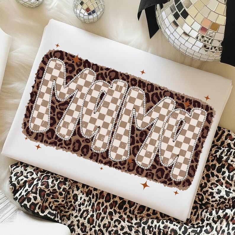 May include: White t-shirt with the word "MAMA" in a brown and tan checkered design, outlined with small silver beads, set against a leopard print background. The shirt is folded, with a leopard print fabric visible.