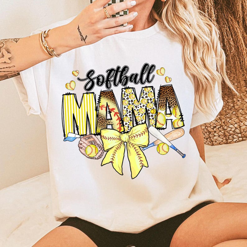 May include: White t-shirt with a graphic design of a softball, a bow, and the text "Softball Mama" in yellow, black, and brown.