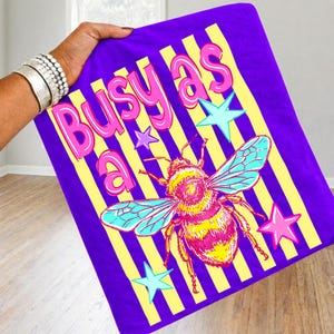 May include: A purple graphic tee with the phrase "Busy as a" in pink letters, yellow stripes, and a colorful bee illustration. The tee also features blue and pink star graphics.