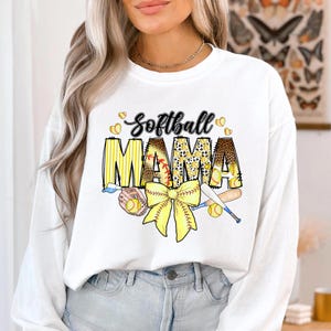 May include: White long-sleeve sweatshirt with the text "Softball Mama" in yellow, black, and brown. The text is surrounded by yellow baseballs and a yellow bow with a baseball bat and glove.