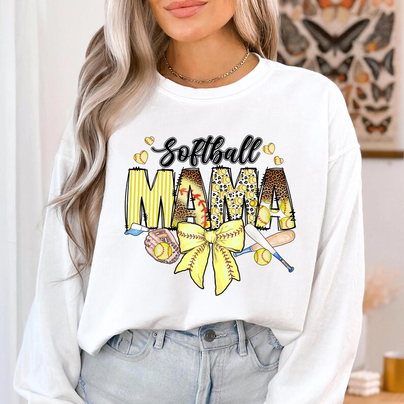 May include: White long-sleeve sweatshirt with the text "Softball Mama" in yellow, black, and brown. The text is surrounded by yellow baseballs and a yellow bow with a baseball bat and glove.