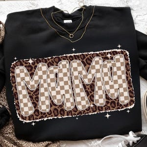 May include: Black sweatshirt with the word "MAMI" in a retro, checkered design. The letters are outlined in white and set against a leopard print background. A gold necklace is visible at the neckline.