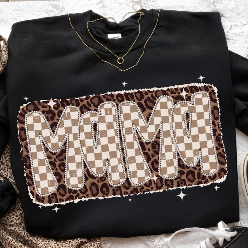 May include: Black sweatshirt with the word "MAMI" in a retro, checkered design. The letters are outlined in white and set against a leopard print background. A gold necklace is visible at the neckline.