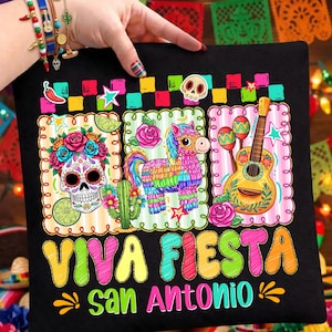 May include: Black t-shirt with a colorful "Viva Fiesta San Antonio" design. The graphic includes a sugar skull, a piñata, and a guitar with maracas. The design is framed with decorative borders and floral elements.