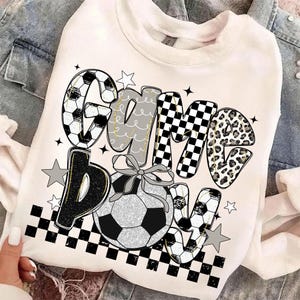 May include: White sweatshirt with the words "Game Bow" in a mix of fonts and patterns, including soccer ball designs, checkered patterns, and leopard print. A soccer ball with a bow is centered below the text, with star accents.