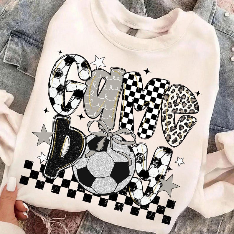 May include: White sweatshirt with the words "Game Bow" in a mix of fonts and patterns, including soccer ball designs, checkered patterns, and leopard print. A soccer ball with a bow is centered below the text, with star accents.