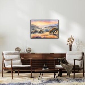 May include: A watercolor painting of a landscape with rolling hills, trees, and a sunset in the background. The painting is framed in a black frame and is hanging on a white wall.