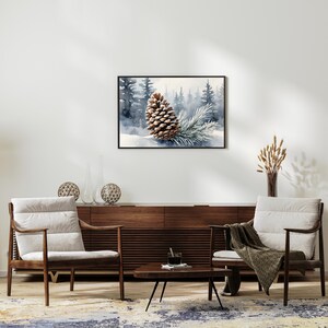 May include: A framed watercolor print featuring a detailed brown pinecone and a sprig of evergreen needles set against a snowy forest backdrop. The artwork is displayed above a mid-century modern style wooden cabinet and two armchairs.
