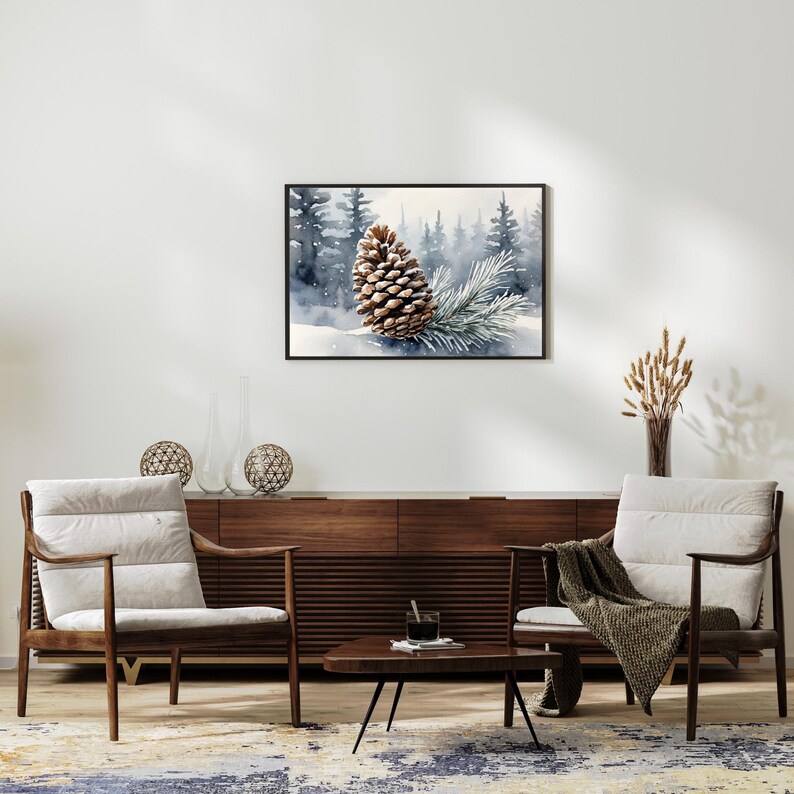May include: A framed watercolor print featuring a detailed brown pinecone and a sprig of evergreen needles set against a snowy forest backdrop. The artwork is displayed above a mid-century modern style wooden cabinet and two armchairs.