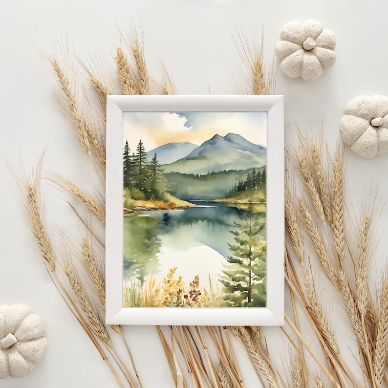 Op de afbeelding: Een ingelijste aquarel schilderij van een serene bergmeersc&egrave;ne. Het kunstwerk, in een witte lijst, toont groene bomen, blauwe bergen en een reflecterende meer. Decoratieve tarwearen en witte pompoenen omringen de lijst.