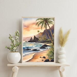 May include: Framed watercolour print depicting a tropical beach with palm trees, mountains, and ocean waves. The artwork is in a light wood frame. The sky displays orange and yellow hues, suggesting a sunset or sunrise over the beach.