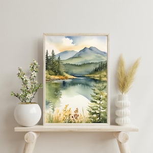 Op de afbeelding: Ingelijste aquarelprint met een serene meerlandschapssc&egrave;ne met bergen op de achtergrond. Het kunstwerk, in een licht houten frame, toont een natuurlijke omgeving met bomen en waterreflecties. Het kleurenpalet omvat groen, blauw en geel.