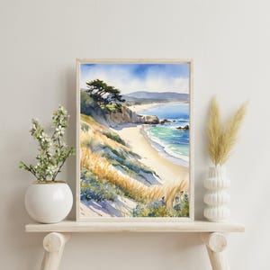 K&ouml;nnte beinhalten: Aquarellgem&auml;lde einer Strandszene mit einem Sandufer, blauem Meer und einem gr&uuml;nen H&uuml;gel mit B&auml;umen. Das Gem&auml;lde ist in einem hellen Holzrahmen gerahmt und auf einem wei&szlig;en Regal ausgestellt.