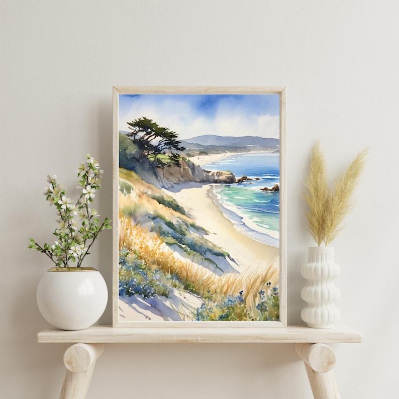 K&ouml;nnte beinhalten: Aquarellgem&auml;lde einer Strandszene mit einem Sandufer, blauem Meer und einem gr&uuml;nen H&uuml;gel mit B&auml;umen. Das Gem&auml;lde ist in einem hellen Holzrahmen gerahmt und auf einem wei&szlig;en Regal ausgestellt.