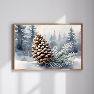 May include: Watercolor painting of a pine cone and evergreen branch in a snowy forest scene. The artwork is framed in a light wood frame. The background features a misty forest with blue and gray tones.
