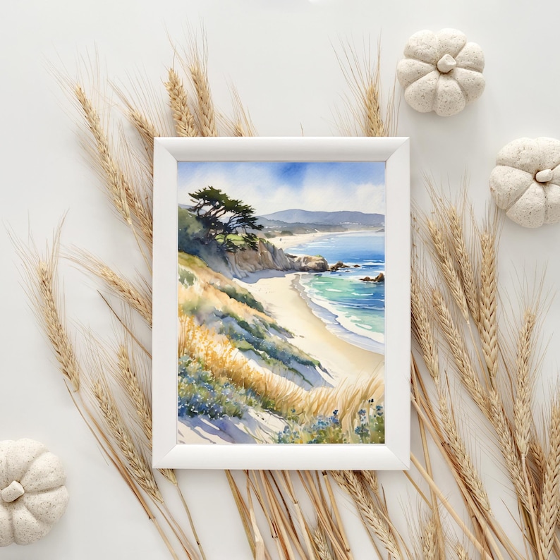 K&ouml;nnte beinhalten: Ein gerahmtes Aquarellbild einer Strandszene mit einer felsigen Klippe, einem Sandstrand und blauem Meerwasser. Das Bild ist von Weizenhalmen und wei&szlig;en K&uuml;rbissen umgeben.