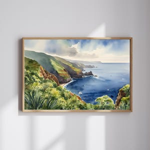 Op de afbeelding: Waterverfschilderij van een kustlandschap met groene heuvels en een diepblauwe oceaan. Een kleine zeilboot is zichtbaar in het water. Het kunstwerk is ingelijst in een lichtbruine houten lijst, wat een serene uitstraling cre&euml;ert.