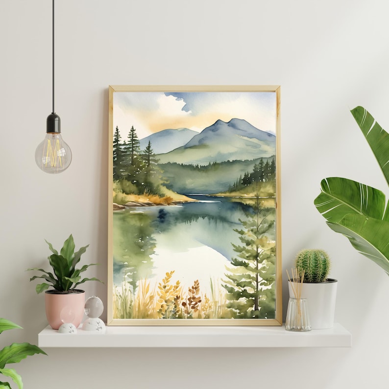 Op de afbeelding: Aquarel schilderij van een serene bergmeersc&egrave;ne. Het kunstwerk heeft een licht houten frame, met een landschap van groene bomen, blauwe bergen en een reflecterend meer. De lucht heeft zachte gele en blauwe tinten.