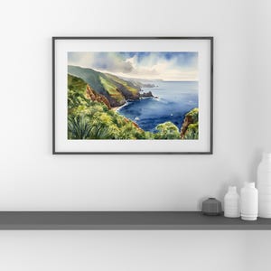 Op de afbeelding: Aquarel schilderij van een kustlandschap met weelderige groene heuvels die de diepblauwe oceaan ontmoeten. Een klein strand en zeilboten zijn zichtbaar. Het kunstwerk is ingelijst in een zwarte lijst en wordt tentoongesteld op een grijze plank met witte vazen.
