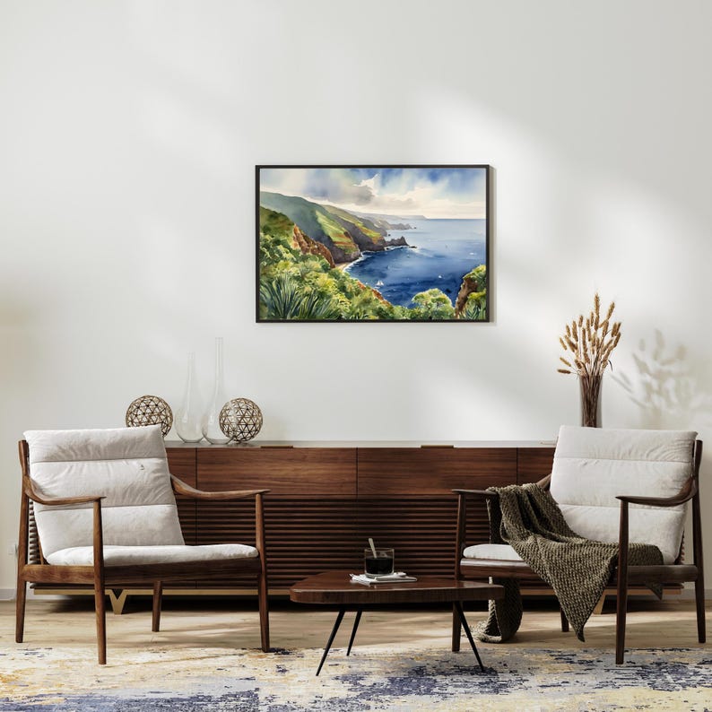 Op de afbeelding: Een ingelijste aquarel van een kustlandschap met kliffen, weelderige groene vegetatie en de blauwe oceaan. Het kunstwerk hangt boven een houten kast in mid-century modern stijl en twee fauteuils met cr&egrave;mekleurige kussens.