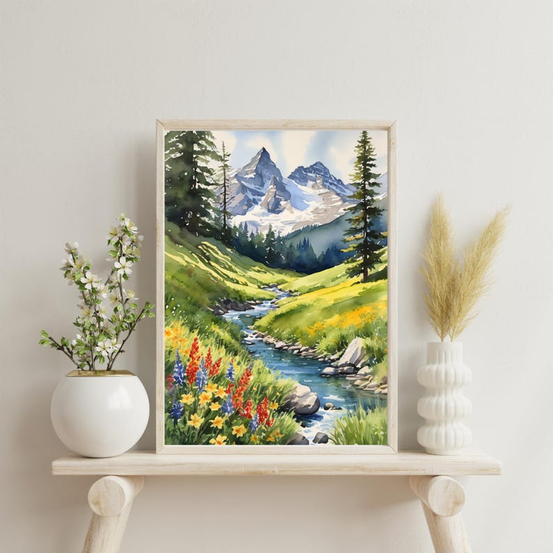 Op de afbeelding: Waterverf schilderij van een berglandschap met een beek die door een vallei stroomt. De bergen zijn met sneeuw bedekt en de vallei is gevuld met groen gras en wilde bloemen. Het schilderij is ingelijst in een licht houten lijst.