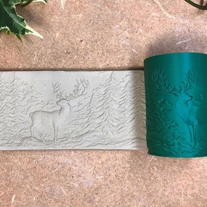 Op de afbeelding: Een groene cilindrische stempel en een rechthoekige stempel met een hert- en bosontwerp. Het hert staat in een bos van bomen. De stempels zijn gemaakt van een hard materiaal. De achtergrond is lichtbruin.