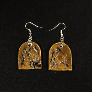 Op de afbeelding: Een paar handgemaakte oorbellen met een botanisch ontwerp. Elke oorbel heeft een bronskleurige, gewelfde hanger met een bloemmotief en zilverkleurige oorhaken. De oorbellen worden gepresenteerd tegen een zwarte achtergrond.