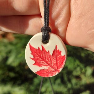 Peut inclure: Un collier avec un pendentif en céramique blanche représentant une feuille d'érable rouge. Le collier est doté d'un cordon noir.