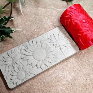 Può includere: Un rullo rosso a motivo di girasoli e un timbro rettangolare con un disegno di girasoli. Il timbro è grigio chiaro e presenta girasoli tridimensionali. Il rullo è rosso e ha un motivo ripetuto di girasoli.