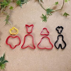 Puede incluir: Una colección de cortadores de galletas coloridos en varias formas, incluyendo una flor amarilla, corazones y campanas rojas, y formas abstractas negras. Estas herramientas para hornear están diseñadas para crear galletas decorativas.