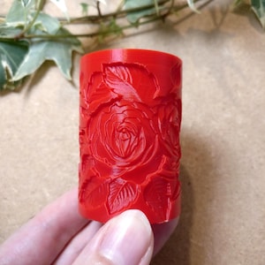 May include: A vibrant red cylindrical object with a detailed rose and leaf pattern carved into its surface. The floral design is prominent, with petals and leaves creating a textured appearance. The object is held in a hand.