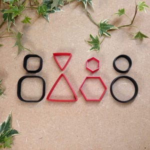 Può includere: Una collezione di formine geometriche per biscotti in varie forme: quadrato, triangolo, esagono e cerchio. Le formine sono rosse e nere, disposte su una superficie marrone chiaro con fogliame verde.