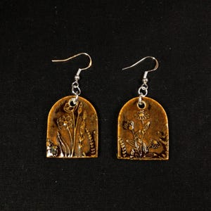 Op de afbeelding: Een paar handgemaakte oorbellen met een botanisch thema. Elke oorbel heeft een bronskleurige, gewelfde hanger met een bloemenontwerp in een lichtere tint. De oorbellen hangen aan zilverkleurige oorhaken.