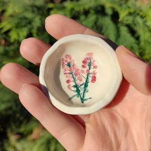 Könnte beinhalten: Eine kleine weiße Keramikschale mit handbemaltem Design aus rosa Blüten mit grünen Stielen.