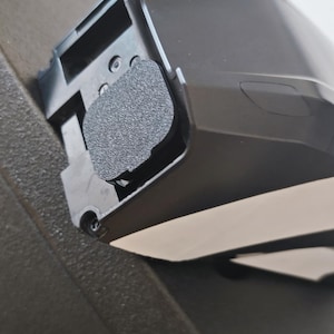 Può includere: Primo piano di un componente automobilistico in plastica nera con un cuscinetto strutturato grigio scuro. Il componente ha una forma curva e sembra far parte di un assemblaggio più grande. Un oggetto rosso è visibile sullo sfondo.